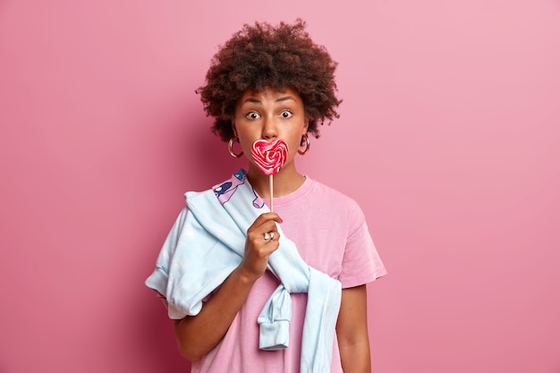 Free photo surprised curly haired woman covers mouth with tasty heart shaped candy lollipop, going to have outdoor walk with boyfriend, dressed in casual t shirt and sweater tied over shoulder. people, youth