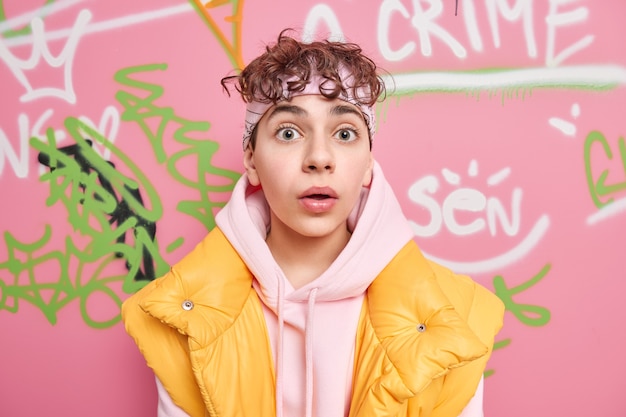 Free photo surprised curly teenage boy looks very shocked at camera hears shocking news wears hoodie with vest holds breath cannot believe his eyes stands at street against graffiti wall