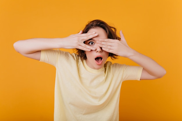 Surprised dark-eyed girl with ring funny posing on orange wall. Pale brunette woman in yellow t-shirt covering face and expressing amazement.