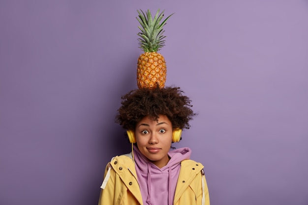 Free photo surprised dark skinned millennial girl carries ripe pineapple on head, listens music, wears headphones on ears, spends free time listening favorite song dressed casually poses indoor. woman with fruit