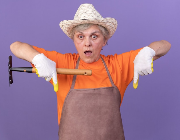 Free Photo surprised elderly female gardener wearing gardening hat and gloves points down holding rake