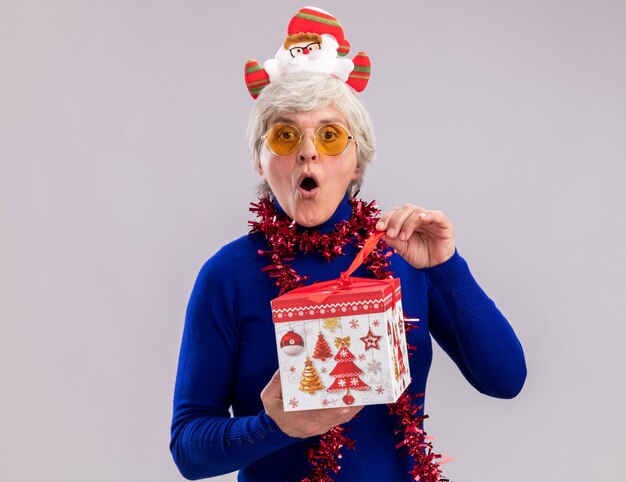 Surprised elderly woman in sun glasses with santa headband and garland around neck opening christmas gift box isolated on white wall with copy space