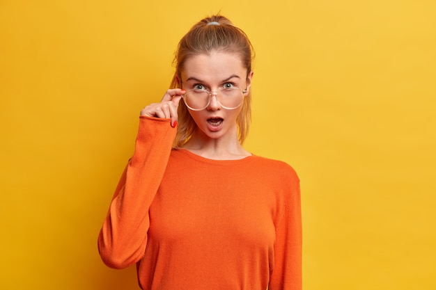 Free photo surprised emotional woman with widely opened mouth looks through optical glasses, dressed in casual orange jumper, hears astonishing news, poses