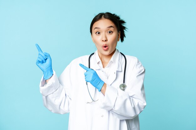 Surprised and enthusiastic asian doctor female medical worker pointing fingers at upper left corner ...