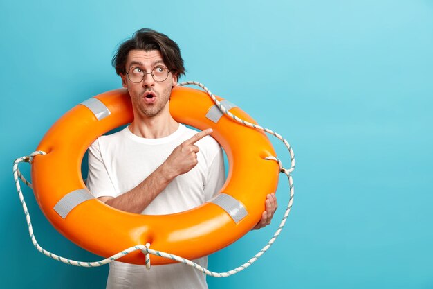 Surprised European man ready to go in sunny place on summer holidays carries inflated lifebuoy points away at blank space