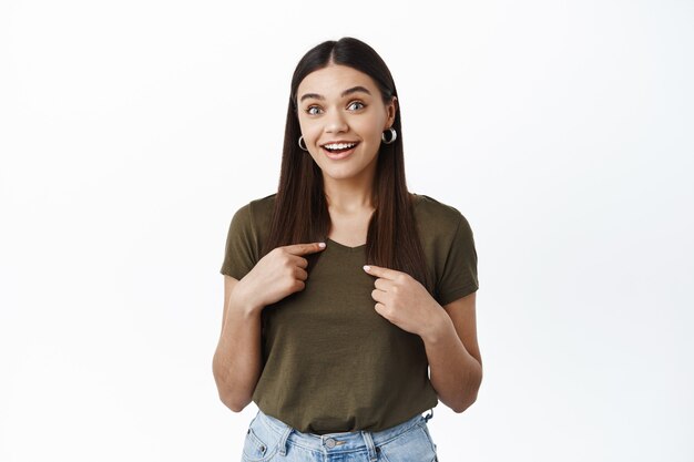 Surprised and excited young woman raising eyebrows from surprise, pointing at herself, being chosen, winning prize, standing against white wall