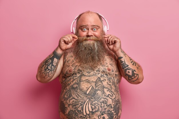  surprised funny chubby man holds mustache, stands shirtless over pink wall, has fat belly, tattoos on stomach and arms, chills with favorite music, wears headphones, enjoys new track