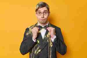 Free photo surprised funny man adjustes bowtie dressed in formal outfit wears round spectacles dresses for special occasion smeared with serpantine spray isolated over yellow background preparing for holiday