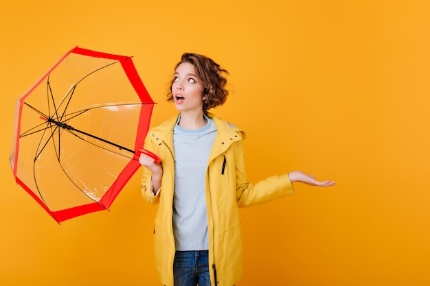 Free photo surprised girl in coat looking up and holding umbrella.  shocked young lady with parasol isolated on bright orange wall.