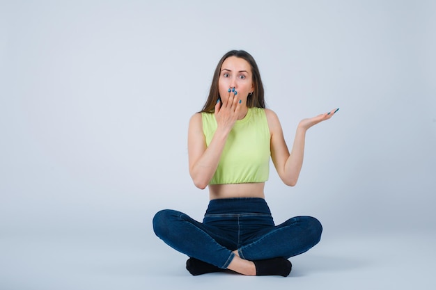 Surprised girl is holding hand on mouth by sitting on floor on white background