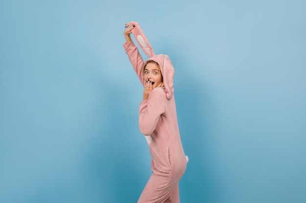 Free photo surprised girl in rabbit costume holding ears on blue background studio shot of pretty young woman wears bunny kigurumi