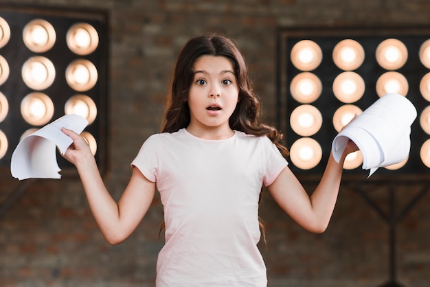 Free photo surprised girl standing in front of stage light holding scripts