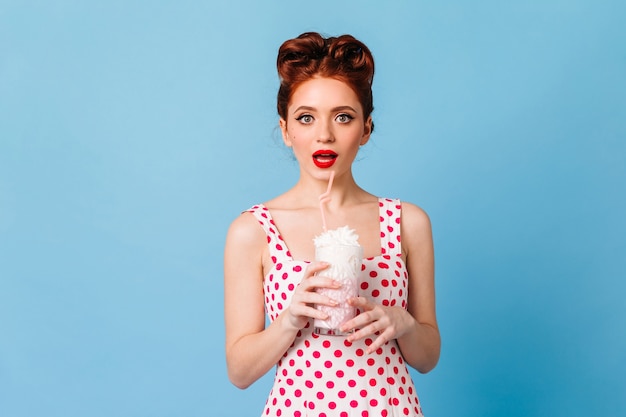 Free photo surprised girl with red lips drinking milkshake. studio shot of emotional young lady in polka-dot dress standing on blue space.