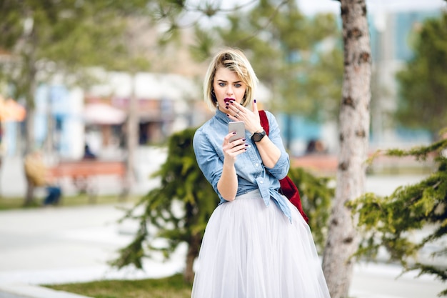 Free photo a surprised girl with short blond hair and bright pink lips looking at a smartphone