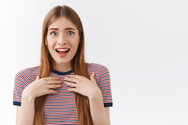 Surprised grateful goodlooking young woman in striped tshirt hold hands on chest impressed and shocked open mouth gasping flattered smiling pleased and delighted white background