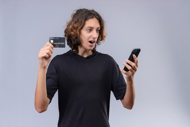 Surprised guy with long hair in black t-shirt holding a phone and a bank card on white background