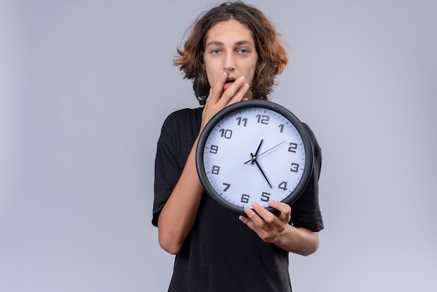 Surprised guy with long hair in black t-shirt holding a wall clock and covered his mouth with hand on white background