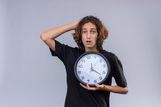 Surprised guy with long hair in black t-shirt holding a wall clock and grabbed his head on white background