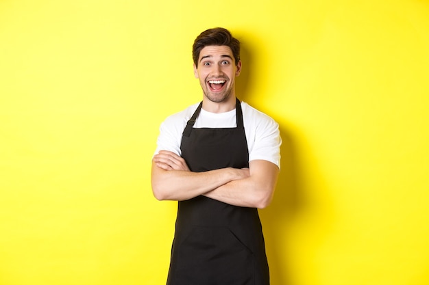 Surprised handsome barista in black apron raising eyebrows, looking amazed, standing against yellow background