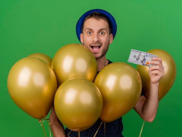 Free Photo surprised handsome caucasian man wearing blue party hat holds helium balloons and money isolated on green background with copy space