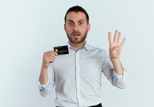 Surprised handsome man holds credit card and gestures four with hand isolated on white wall