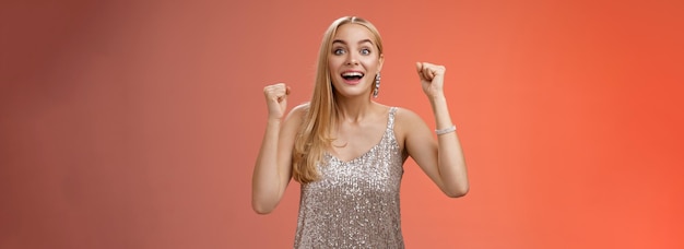 Free photo surprised happy celebrating blond young woman in silver trendy dress raising hands up yes victory gesture smiling broadly excited winning first place reach goal grinning thrilled triumphing