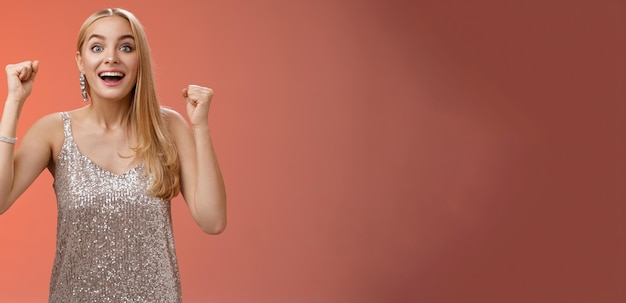 Surprised happy celebrating blond young woman in silver trendy dress raising hands up yes victory