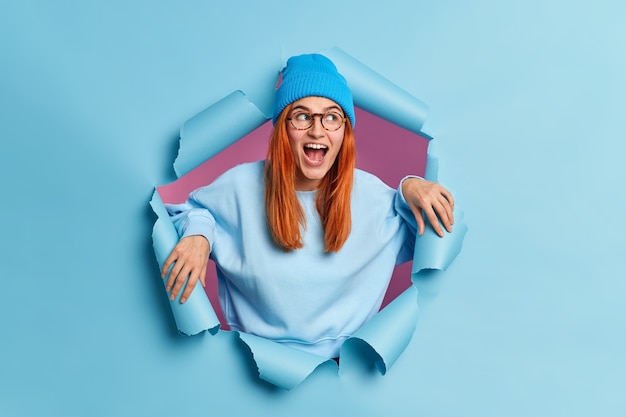 Surprised happy woman looks with great interest aside keeps mouth opened wears blue hat and sweatshirt breaks through paper hole