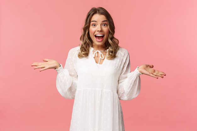 Surprised and happy young blond girl spread hands sideways smiling, standing pink wall lucky and upbeat.