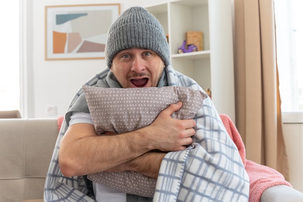 surprised ill man with scarf around neck wearing winter hat wrapped in plaid holding pillow sitting on couch at living room