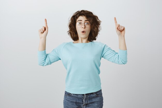 Surprised and impressed woman in glasses showing something awesome, pointing fingers up