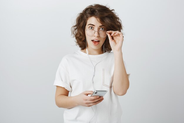 Surprised and interested young woman looking excited while using earphones and mobile phone