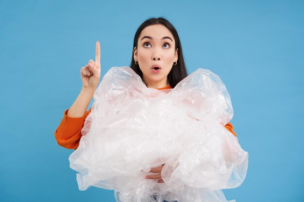 Surprised korean woman with plastic waste points finger up and looks on top blue background