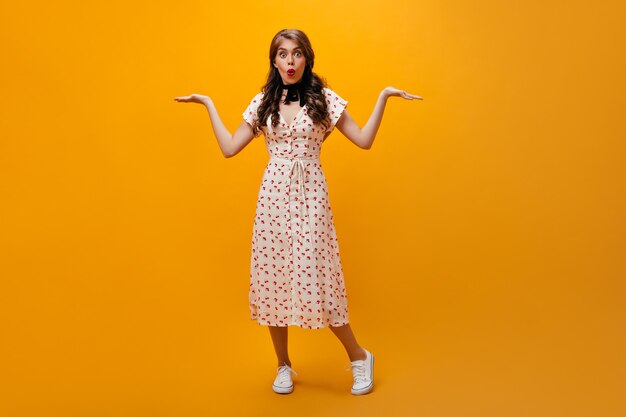 Surprised lady shrugging on orange background.Trendy young woman with wavy hair in long white dress looking into camera.