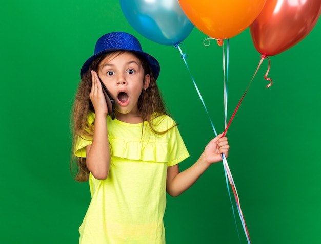 surprised little caucasian girl with blue party hat holding helium balloons and talking on phone isolated on green wall with copy space