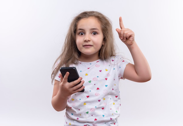 Surprised little school girl wearing white t-shirt holding phone points to up on isolated white background