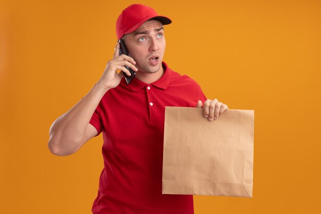 Surprised looking at side young delivery man wearing uniform with cap holding paper food package speaks on phone isolated on orange wall