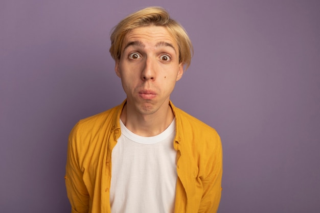 Free photo surprised looking straight ahead young blonde guy wearing yellow t-shirt holding hands on waist isolated on purple