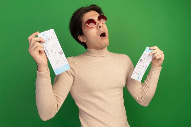 Free photo surprised looking up young handsome guy wearing glasses holding tickets isolated on green wall