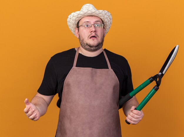 Surprised looking up young male gardener wearing gardening hat and gloves holding clippers isolated on orange wall