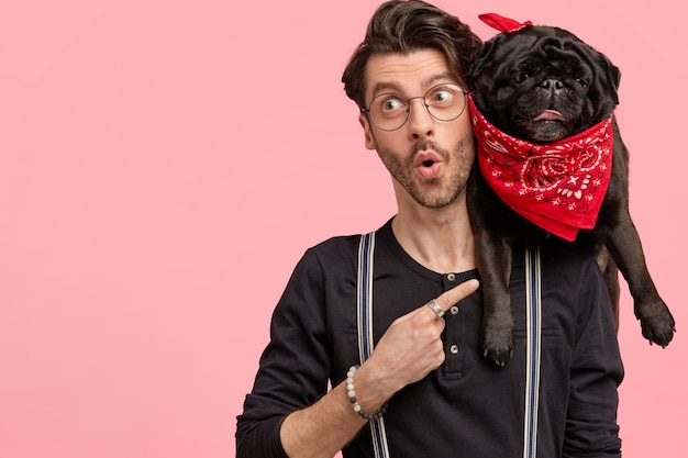 Free Photo surprised male hipster dressed in fashionable black shirt, points at his funny dog with red bandana, feels surprised as bought it for low price, isolated over pink wall with blank space on left