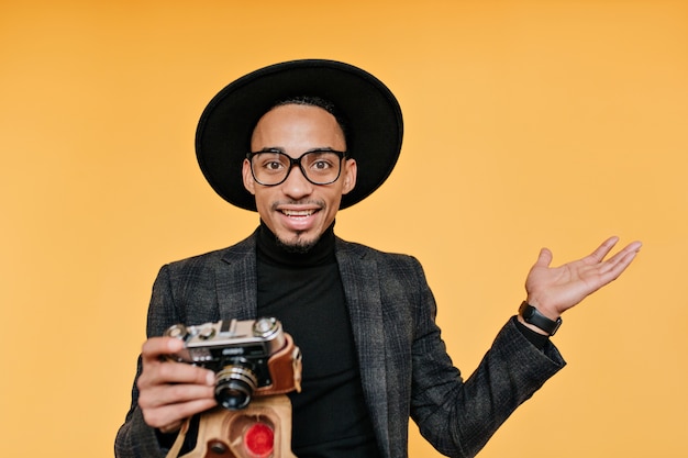 Free photo surprised male photographer in black hat. indoor photo of african young man with camera isolated on yellow wall.