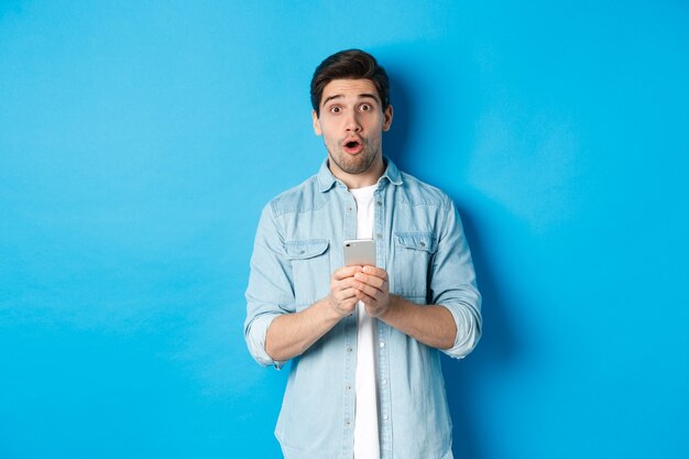 Surprised man in casual clothes looking astounded, holding smartphone, standing against blue background.