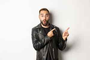 Free photo surprised man in cool black leather jacket pointing fingers at upper right corner, showing logo or banner, white background.