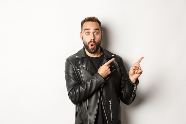 Surprised man in cool black leather jacket pointing fingers at upper right corner, showing logo or banner, white background.