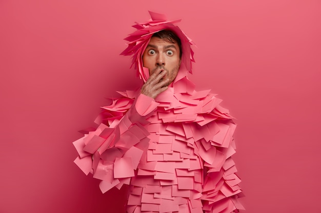 Free photo surprised man covers mouth and stares with bugged eyes, afraids of something, hears astonishing news, gasps from shocking rumors, wears adhesive notes, isolated on pink wall. omg concept