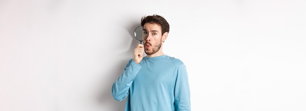 Free photo surprised man found something looking through magnifying glass with amazed face standing over white