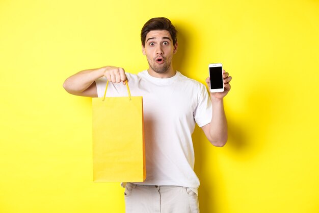 Surprised man holding shopping bag and showing smartphone screen, concept of mobile banking and app achievements, yellow background.