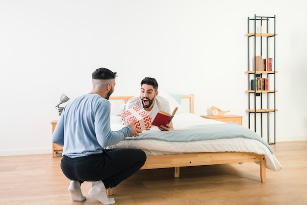 Free photo surprised man lying on bed holding book in hand looking at his boyfriend giving gift box