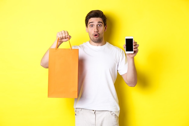 Free Photo surprised man showing mobile screen and shopping bag, standing against yellow background. copy space